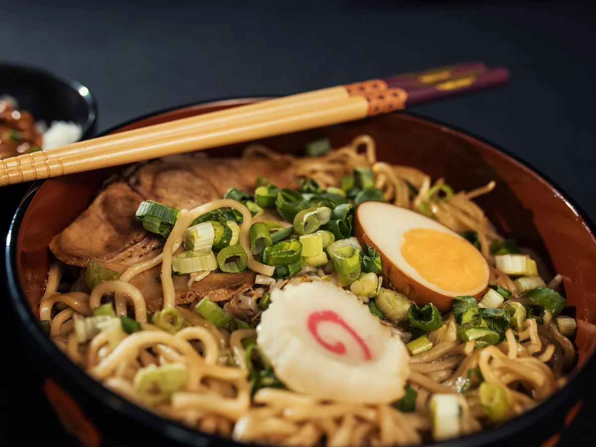 Close view of a bowl of ramen with chashu pork, a soft-boiled egg half, a pink-swirled fish cake and chopped spring onion under wooden chopsticks